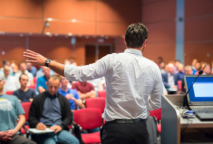 Mężczyzna podczas wykładu na ScalaSphere DevTools Summit. W tle siedzą notujący mężczyźni