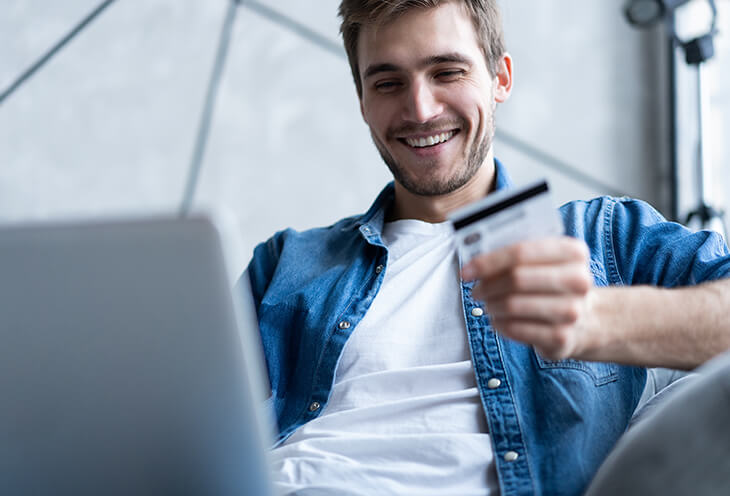 Młody uśmiechnięty mężczyzna dokonuje płatności na laptopie i w ręce trzyma kartę kredytową 