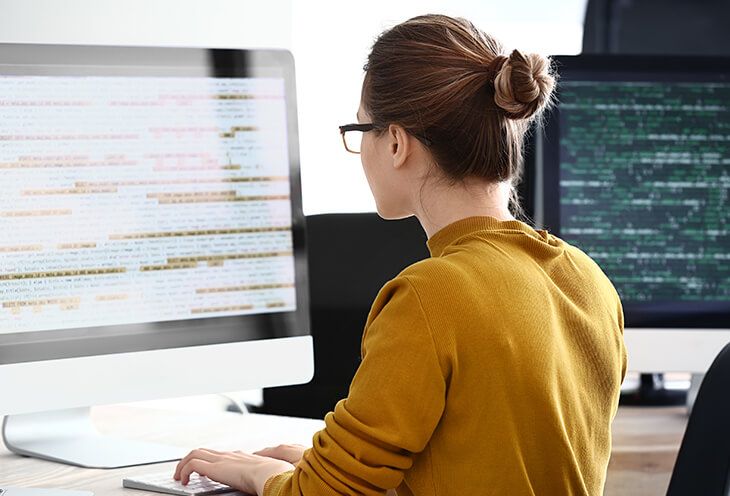 Młoda kobieta w żółtym swetrze uczy się programowania pracując na dużym monitorze 