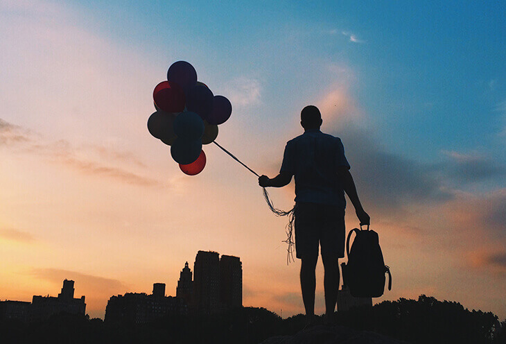 Młody mężczyzna stoi w cieniu. W ręce trzyma balony i plecak