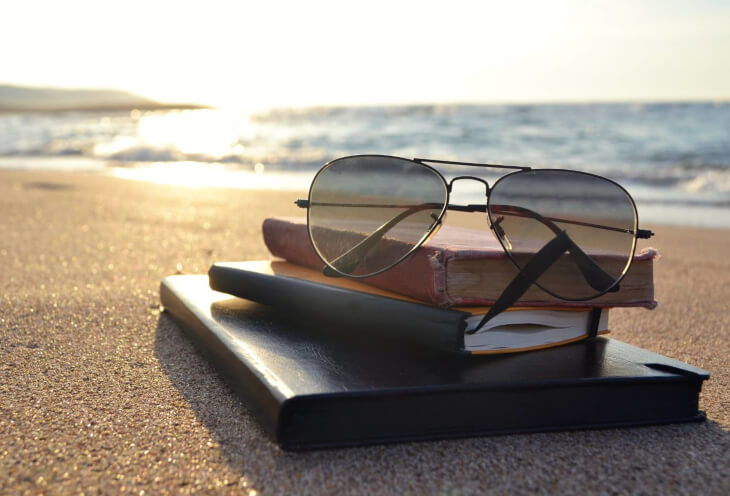 Jak wygląda odpoczynek od pracy? Okulary leżą na książkach na plaży. W tle widać może i słońce