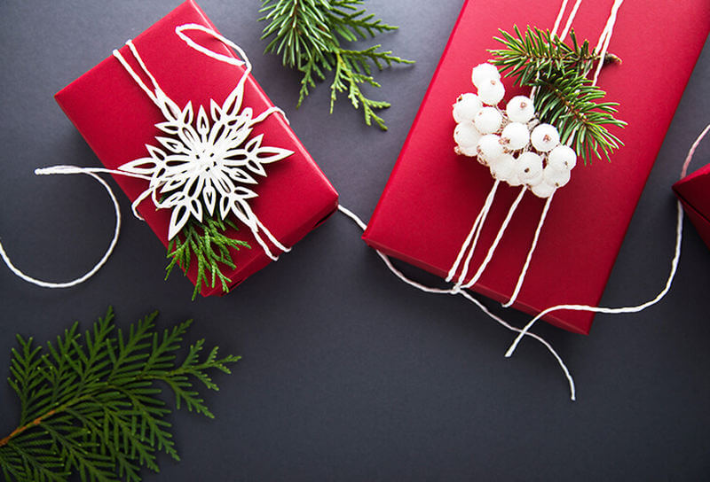 Dwa  bożonarodzeniowe prezenty udekorowane białą wstążką. Wokół porozrzucana jest choinka
