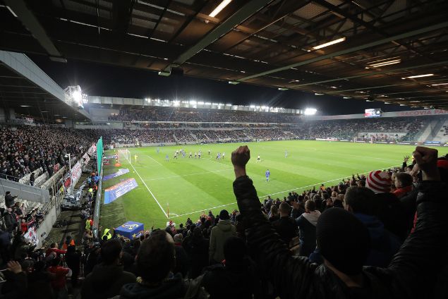 Trybuna stadionu klubu Cracovia. Ręka wystająca w tłumie kibiców na znak zwycięstwa.