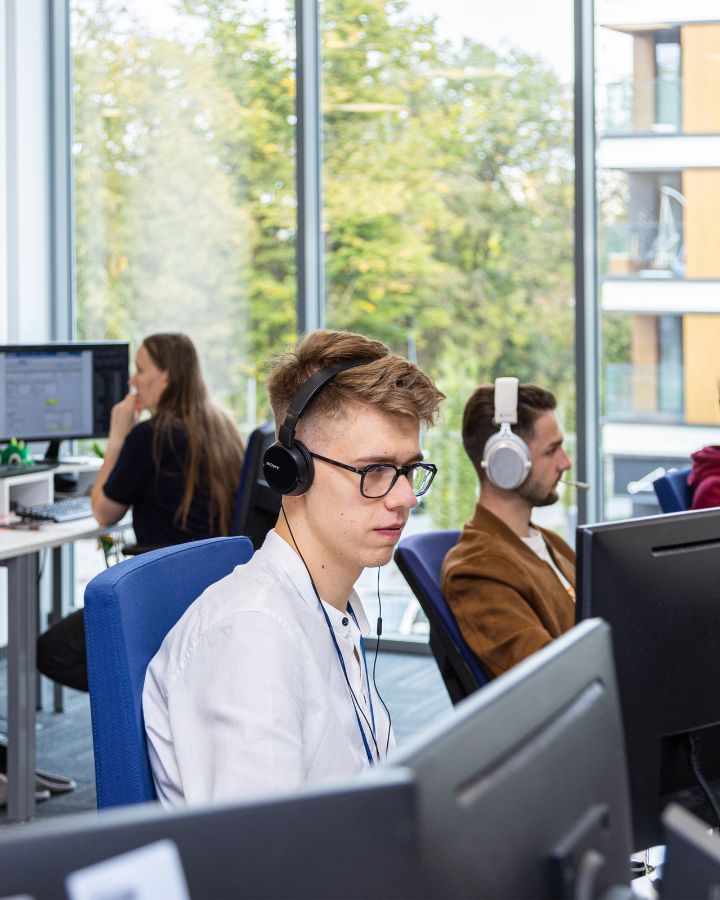Na pierwszym planie młody mężczyzna podczas pracy przy komputerze, w tle jeszcze inne osoby i zieleń za oknem