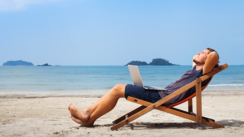 Mężczyzna odpoczywający na leżaku z przenośnym komputerem na kolanach. W tle morska plaża.