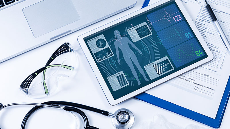 Na biurku u lekarza leży laptop, tablet, dokumenty medyczne okulary oraz stetoskop