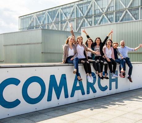 Grupa pracowników  siedząca na murku z dużym logo Comarch, patrzących w kierunku obiektywu, z uniesionymi rękami.